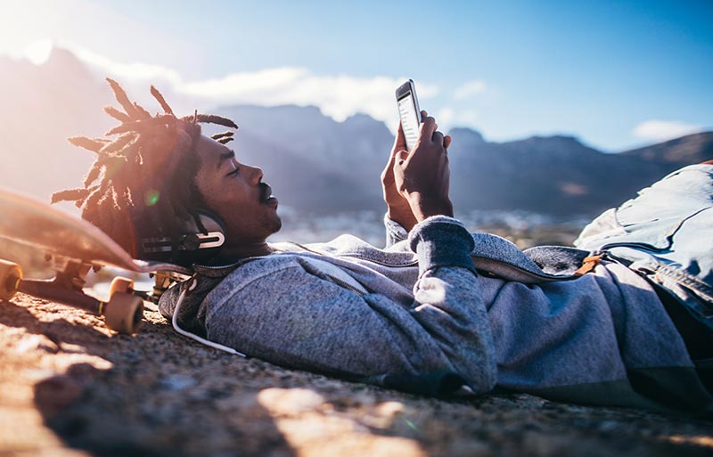Close up shot of a man lying on ground with head propped up on skateboard while looking at his mobile device. We see mountains in the background.