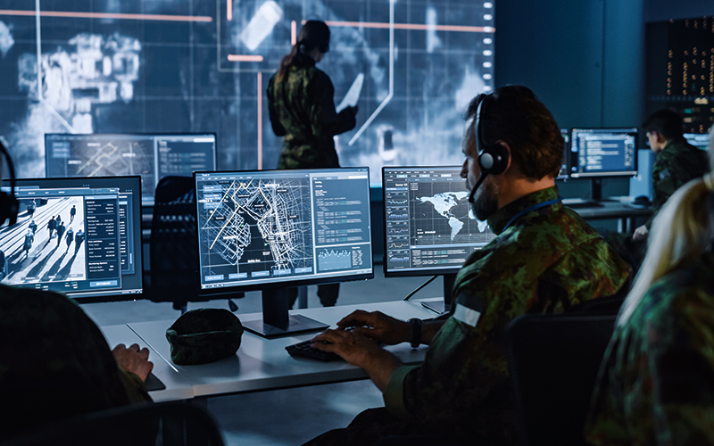 Man in military attire sitting in front of several monitors in a control room.
