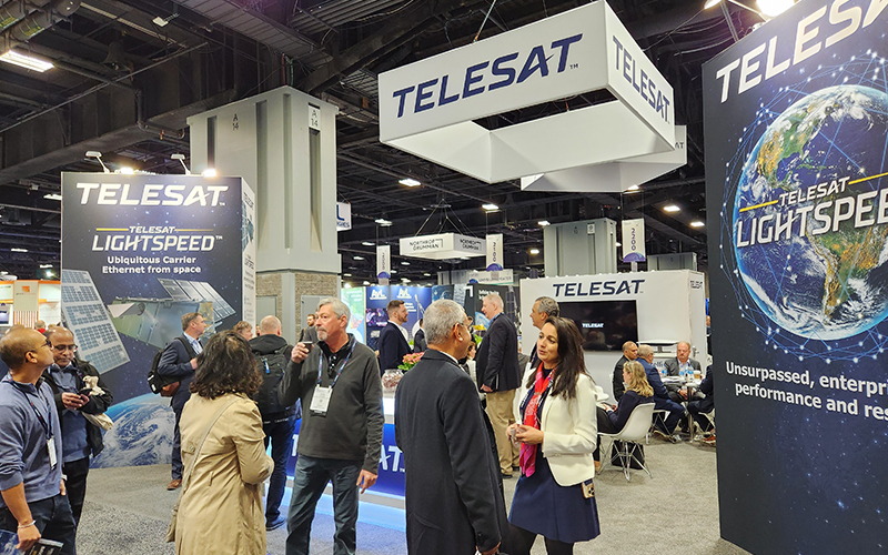 Attendees interact and walk through a bustling tradeshow floor, with groups of people networking and exploring exhibits. In the background, the Telesat booth stands prominently with branded signage, display monitors, and promotional materials visible, drawing the attention of several visitors.