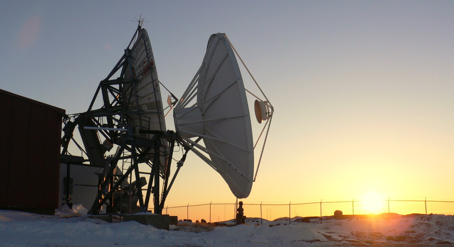 Large satellite ground station antennas in a snowy landscape at sunset, silhouetted against the sky, representing remote communications infrastructure in a northern environment