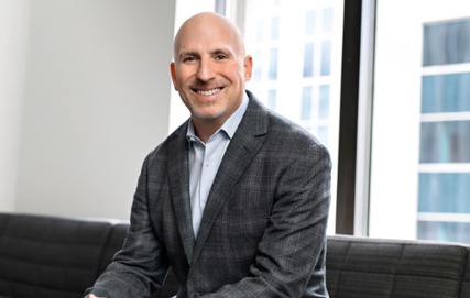 Glenn Katz of Telesat in a grey blazer seated and smiling in a modern office with city windows in the background