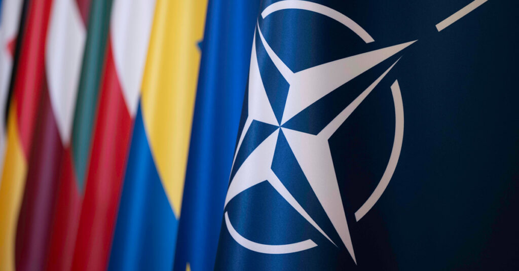 Close-up of the NATO flag showing the white compass rose emblem on a dark blue background, with blurred national flags of member countries visible behind it.