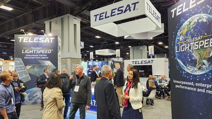 Attendees interact and walk through a bustling tradeshow floor, with groups of people networking and exploring exhibits. In the background, the Telesat booth stands prominently with branded signage, display monitors, and promotional materials visible, drawing the attention of several visitors.