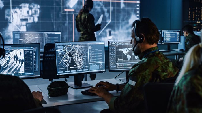 Man in military attire sitting in front of several monitors in a control room.