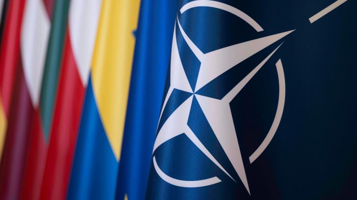 Close-up of the NATO flag showing the white compass rose emblem on a dark blue background, with blurred national flags of member countries visible behind it.