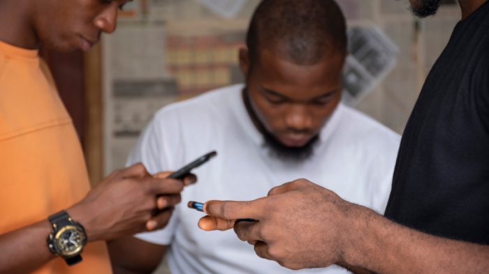 Three young African people sit together, focused on their mobile phones and laptops, collaborating and sharing digital content.